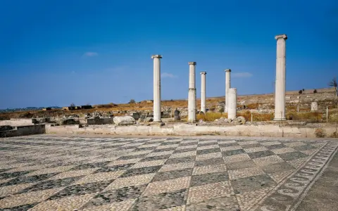 Ruines antiques avec colonnes imposantes et un sol en mosaïque de galets, évoquant l'origine d'Alexandre le Grand.