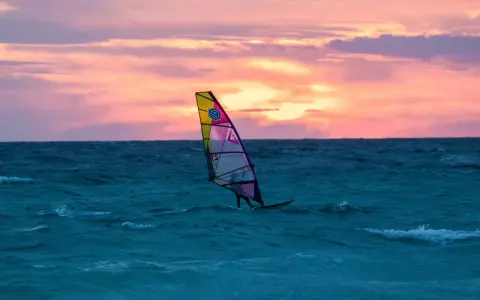 Ein Windsurfer gleitet bei Sonnenuntergang über das Meer. Die bunten Wolken spiegeln sich im Wasser. Ein perfekter Tag für Windsurfen Mallorca.