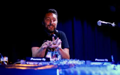 Un dj hip hop souriant, mains en l'air, mixe sur scène devant un public invisible.