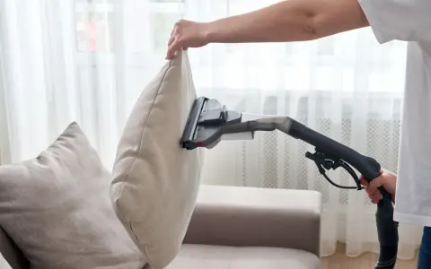 En person använder en soffdammsugare för att rengöra en ljusbeige kudde på en soffa.