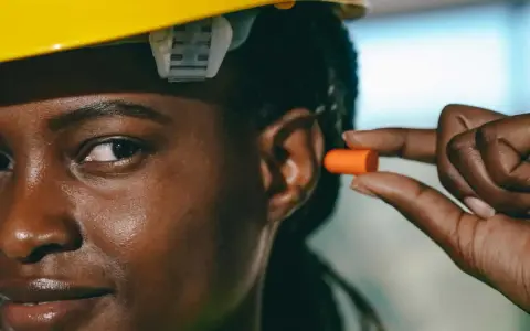 Une femme porte un casque jaune et s'apprête à insérer un bouchon d'oreille orange pour éviter le sifflement.