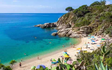 Sonniger Strand in der Toskana mit türkisfarbenem Wasser, Felsen und vielen bunten Sonnenschirmen.