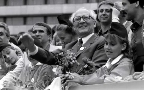 Ein Mann mit Brille winkt lächelnd, umgeben von Menschen. War die DDR eine Diktatur? Diese Szene zeigt eine öffentliche Veranstaltung, vielleicht ein Zeichen der Hoffnung.