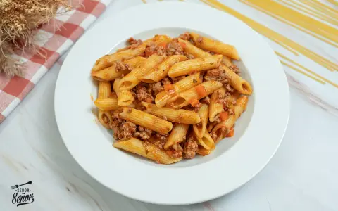 Plato de macarrones con carne picada y salsa de tomate, decorado con un paño a cuadros y espaguetis secos al fondo.