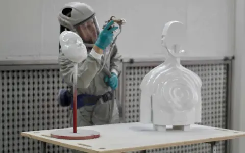 A person in a protective suit sprays a white 3D printed bust and torso, demonstrating how to paint 3d prints for a smooth finish.