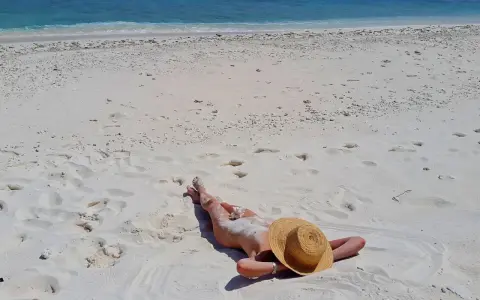 Słoneczna plaża, błękitne morze i osoba opalająca się na piasku. W tle widać fale i biały piasek z licznymi śladami stóp.