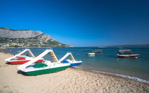 Plaża z łódkami i zjeżdżalniami, idealna na wakacje w camping Galeb. W tle góry i turkusowe morze.