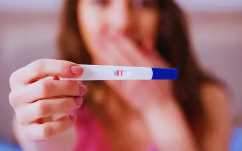 Dwa czerwone paski na teście ciążowym. Kobieta z zakrytymi ustami, być może planuje zwolnienie lekarskie w ciąży.
