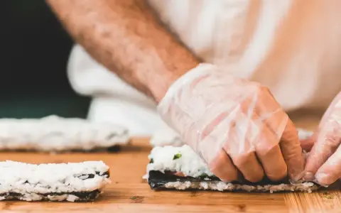 Ręce w rękawiczkach zwijają sushi. Na desce leżą gotowe rolki i składniki do kolejnej. Idealna zalewa do sushi podkreśli smak.