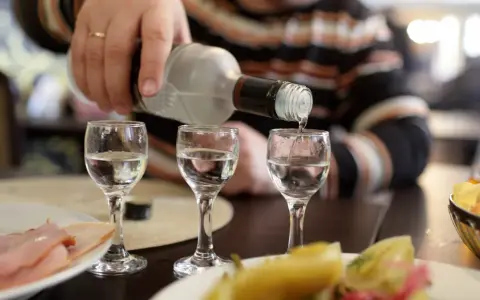 Ręka nalewa wódkę do kieliszków, przygotowując się na rekord promili. Na stole wędlina i przekąski.