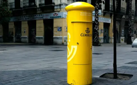 Jasnożółta skrzynka pocztowa Correos, symbol poczty Hiszpanii, stoi na brukowanym placu przed budynkami.