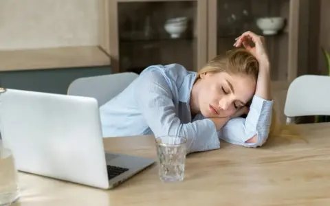 Zmęczona kobieta śpi na biurku obok laptopa. Objawy wypalenia zawodowego, czas na L4.