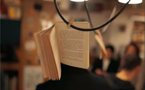 Un libro aperto, appeso per creare una scena da foto libri aesthetic. Le pagine sono piene di testo, con persone sfocate sullo sfondo.