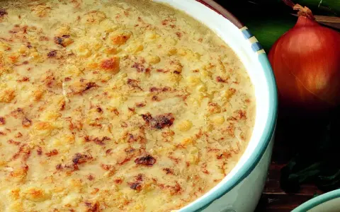 Una ricca zuppa di cipolle gratinata, con crostini dorati e formaggio fuso. Accanto, una cipolla rossa e foglie verdi.