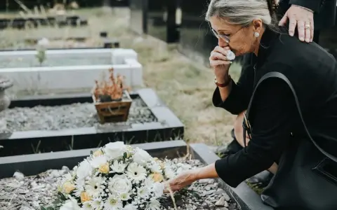 Kobieta w żałobie klęczy przy grobie, ocierając łzy. Bukiet kwiatów leży na ziemi. To smutne ostatnie pożegnanie.