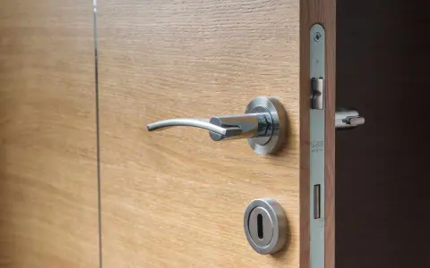 Maniglia e serratura di una porta in legno chiaro, con un'ombra scura che suggerisce una porta che cigola.