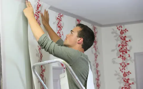 Mann tapeziert mit Vliestapete eine Wand mit rotem Blumenmuster.