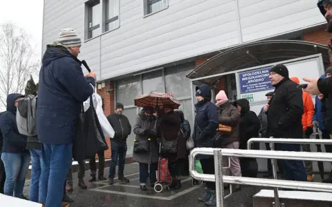Grupa ludzi stoi przed budynkiem spółdzielni mieszkaniowej w deszczu, niektórzy pod parasolem.