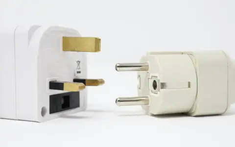Adaptadores para diferentes tipos de enchufes: uno blanco con tres clavijas doradas y otro beige con dos clavijas plateadas.