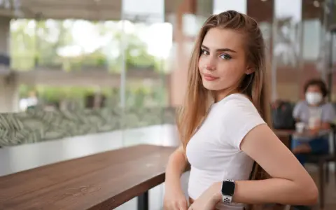 Uśmiechnięta blondynka w białej bluzce, z zegarkiem na ręku, siedzi przy stoliku. Czy to jej sekret, jak uwieść faceta?