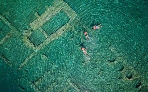Swimmers explore the sunken city of Epidaurus, its ancient stone structures visible beneath the clear turquoise water.