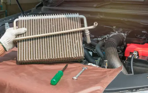 Gloved hand holds car AC evaporator. Tools and engine parts visible. Learn how to make AC colder in car by fixing this component.