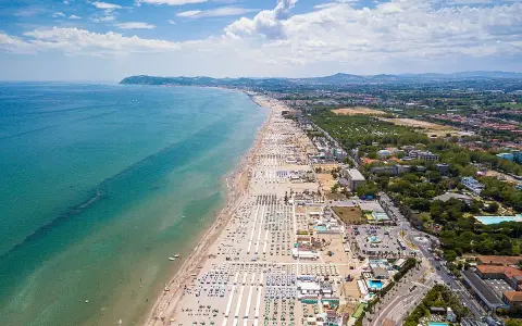 Vista aerea della Riviera Romagnola con spiaggia affollata, mare azzurro e verdeggiante entroterra.