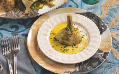 Un carciofo alla romana, tenero e profumato, adagiato su un piatto bianco con olio dorato e prezzemolo tritato.