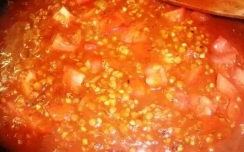 Buchweizen kochen mit Tomaten. Ein Holzlöffel rührt in der roten Sauce mit den Buchweizenkörnern und Tomatenstücken.