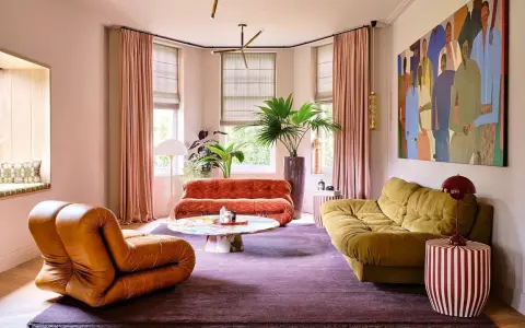 Comment amenager un salon : un fauteuil orange, un canapé rouille, un canapé moutarde, une table basse colorée, un tapis violet et une grande peinture.