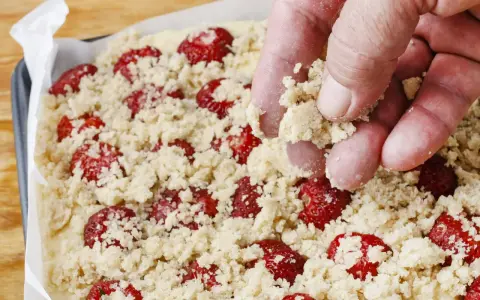 Ręka posypuje kruszonką ciasto z truskawkami. Zapowiada się pyszne ciasto z truskawkami i kruszonką!