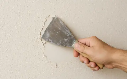 Mano con espátula rasca gotele de una pared, eliminando la textura irregular para prepararla.