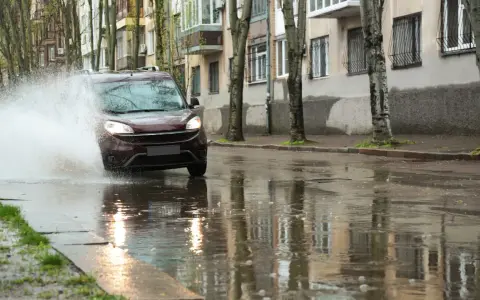 Ciemny samochód wpada w kałużę, powodując potężny **aquaplaning**. Woda rozpryskuje się na boki, a odbicia budynków i drzew tańczą na mokrej nawierzchni.