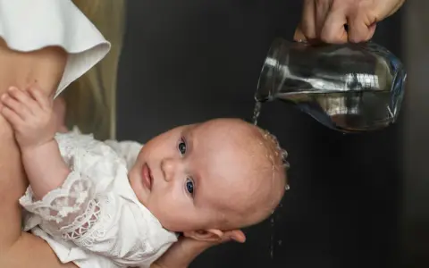 Chrzest święty. Woda spływa na główkę dziecka w białej szatce. Piękne życzenia na chrzest dla maluszka.