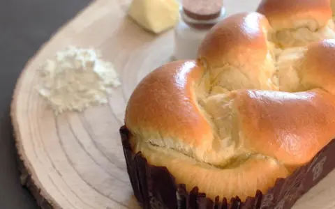 Un pan brioche dorado y esponjoso, listo para ser disfrutado. A su lado, ingredientes para su elaboración: harina, mantequilla y sal.