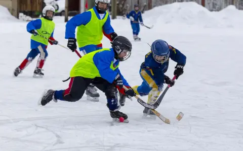 Barn i full fart med hockeyklubbor och hjälmar, ivriga att vinna pucken. De har roligt med lekar på isen.
