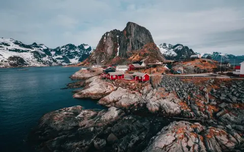 Röda fiskestugor vid en klippig kust med snöklädda berg i bakgrunden. En av de fina platserna i Norge.