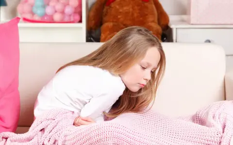 Dziewczynka z bólem brzucha, przykryta różowym kocem, może mieć zatrucie pokarmowe u dziecka.