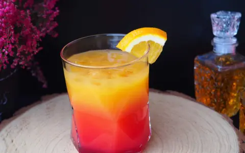 Kolorowy drink z koniakiem, ozdobiony plasterkiem pomarańczy, na tle karafki z bursztynowym alkoholem i kwitnących wrzosów.