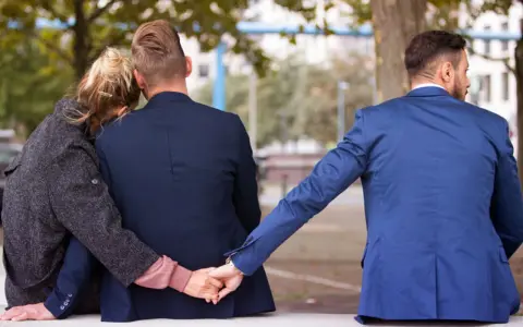 Trójkąt miłosny. Dwoje ludzi siedzi blisko siebie, a trzeci mężczyzna dyskretnie trzyma ich za rękę. To pokazuje, dlaczego zdrada emocjonalna tak boli.