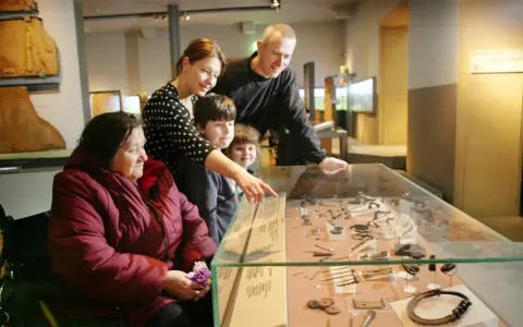 A family, including a woman in a wheelchair, examines artifacts in a museum display, perhaps inspired by famous art curators.