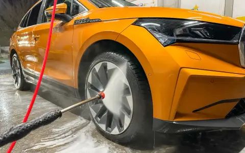 Pomarańczowe auto w trakcie mycia. Woda pod ciśnieniem spływa po karoserii i felgach, tworząc pianę.