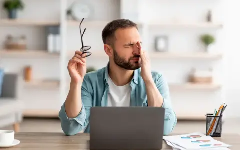 Homme fatigué, se frottant les yeux, lunettes à la main, devant son ordinateur. Il souffre de mal aux yeux à cause des écrans.