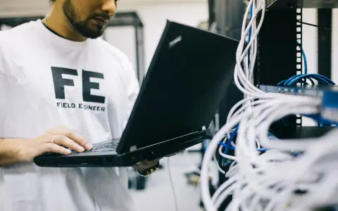 Inżynier w koszulce "FE FIELD ENGINEER" używa laptopa do sprawdzenia **trace route cmd** w serwerowni z plątaniną kabli.