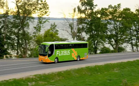Zielony autobus Flixbus jedzie drogą przy jeziorze otoczonym drzewami.