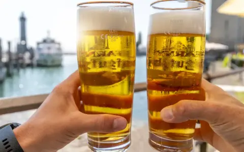 Zwei Hände halten Biergläser mit schaumigem Bier. Im Hintergrund ein Hafen. Ist Bier gesund? Ein Genuss am Wasser.