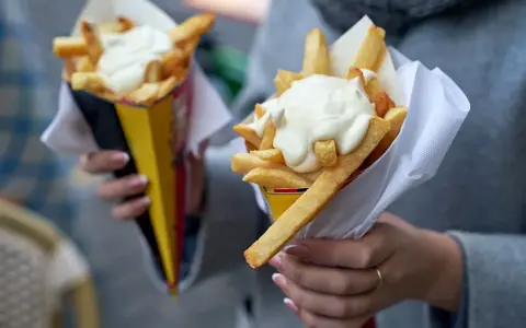 Dwie porcje frytek z majonezem w papierowych opakowaniach, trzymane w dłoniach.