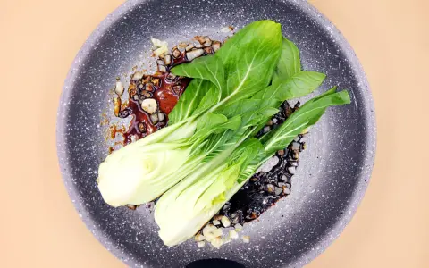 Na patelni smaży się aromatyczny sos z czosnkiem i cebulą, a na nim leży świeża sałata pak choi, gotowa do przygotowania pysznego dania.