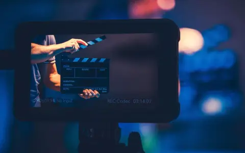 Ręka zamyka klaps filmowy. Na ekranie widać napis "REC:Codec 03:14:07". To plan średni, zapowiadający rozpoczęcie nagrania.