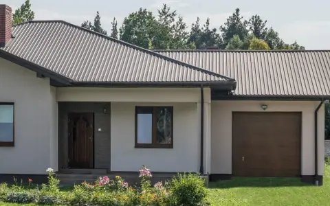 Dom z dachem z blachy trapezowej. Wymiary dachu dopasowane do nowoczesnej bryły budynku. Przed domem ogród z kwiatami.